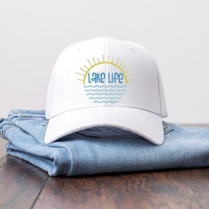 May include: A white baseball cap featuring a graphic design of a yellow sun rising over blue wavy lines with the text Lake Life in blue script lettering. The cap is constructed with a structured crown and a curved brim, showcasing a clean aesthetic.