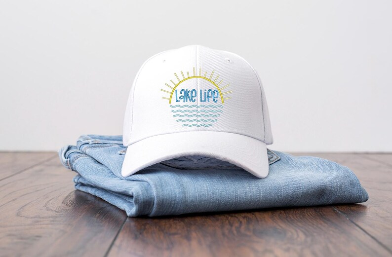 May include: A white baseball cap featuring a graphic design of a yellow sun rising over blue wavy lines with the text Lake Life in blue script lettering. The cap is constructed with a structured crown and a curved brim, showcasing a clean aesthetic.