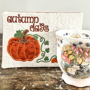 May include: A rectangular white quilted fabric mat featuring an embroidered orange pumpkin with green vines and the text "autumn days" in brown, positioned next to a white ceramic mug decorated with a colourful botanical and berry illustration.