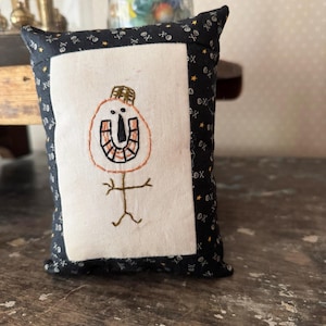 May include: Small, rectangular decorative pillow with a Halloween theme. The pillow features a white center with an embroidered orange and brown pumpkin-like face and stick figure body. The border is black with white skulls and stars.