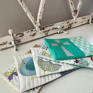 May include: A stack of four quilted fabric coasters with embroidered designs. The top coaster features a turquoise cross, the second coaster has a green umbrella, the third coaster has a blue bird, and the bottom coaster has a butterfly.