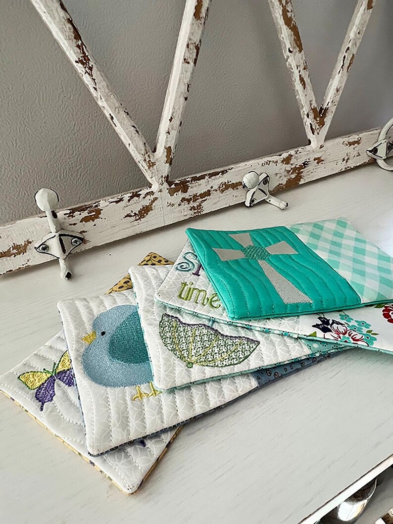 May include: A stack of four quilted fabric coasters with embroidered designs. The top coaster features a turquoise cross, the second coaster has a green umbrella, the third coaster has a blue bird, and the bottom coaster has a butterfly.