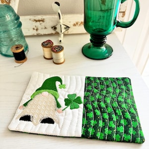 May include: A quilted St. Patrick's Day coaster featuring a gnome with a green hat and a shamrock. The coaster has a white section with the gnome and a green shamrock, and a green shamrock pattern section. Other items include a green glass and spools of thread.