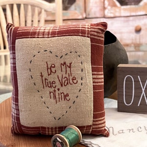 May include: A small, red and white plaid pillow with a heart-shaped design and the text "Be my true Valentine" embroidered on it. The pillow is sitting on a wooden surface with a spool of green thread.
