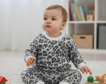 May include: A baby wearing a gray and white leopard print pajama set sits on a white rug. The baby is surrounded by colorful wooden blocks and toys. A mobile with colorful shapes hangs above.