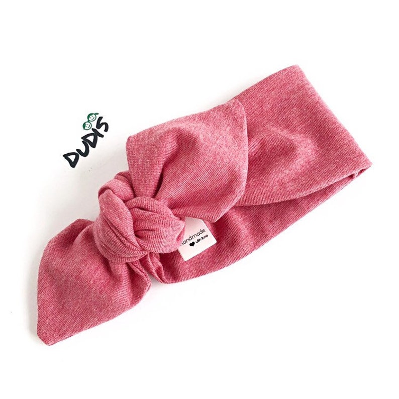 Puede incluir: Una diadema rosa anudada con una peque&ntilde;a etiqueta blanca que dice "hecho a mano con amor".