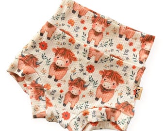 May include: Cream-colored baby shorts with a repeating pattern of cartoon Highland cows, orange flowers, and green foliage. The shorts have a ribbed texture and a wide waistband. A small tag is visible on the side.