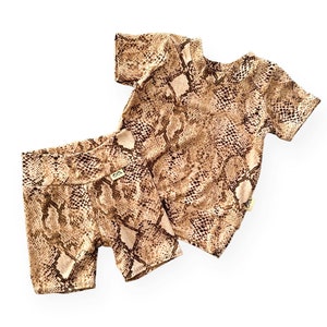 May include: A two-piece outfit for babies and toddlers, consisting of a short-sleeved t-shirt and shorts. Both pieces are made from a soft, stretchy fabric with a brown and beige snake print.