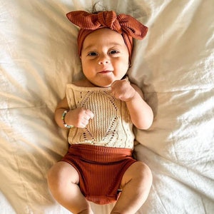 Könnte beinhalten: Ein Baby trägt ein rostiges Stirnband, ein cremefarbenes Häkel-Top und eine rostige Hose. Das Baby liegt auf einem weißen Laken.