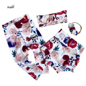 May include: A baby outfit set with a white base and a floral pattern in shades of pink, red, and purple. The set includes leggings, a headband, and a diaper cover.