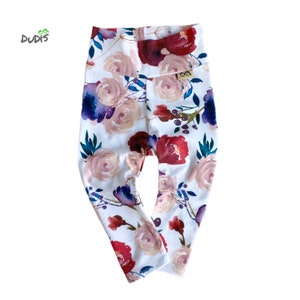 May include: White leggings with a floral print featuring pink, red, and blue watercolor flowers. The leggings have a high waist and a small brown label with the text "OOA" on the right side.