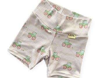 May include: Tan ribbed shorts with a repeating pattern of green tractors with pink wheels. The shorts have a wide waistband and a small yellow tag with a logo. The shorts are made of a soft, stretchy material.