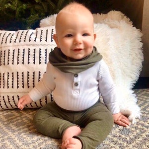 May include: A baby wearing a white ribbed long-sleeve shirt with a single button closure, olive green pants, and a matching olive green scarf. The baby is sitting on a white fluffy rug.