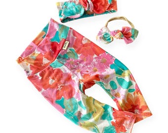 May include: A floral print baby leggings set with a matching headband and bow. The leggings, headband, and bow feature a vibrant pattern of pink, orange, and teal flowers on a white background. The set is designed for infants.