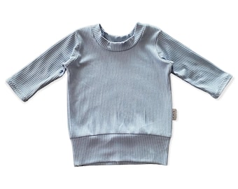 Puede incluir: Una camiseta de manga larga acanalada azul claro. La camiseta tiene cuello redondo y dobladillo acanalado. La tela tiene una textura acanalada vertical. Una pequeña etiqueta es visible en la parte inferior derecha.
