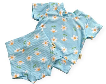 May include: A light blue, ribbed knit two-piece set featuring a daisy pattern. The set includes a short-sleeved top and high-waisted shorts. The daisies have white petals and orange centers with green leaves. The set is designed for babies or toddlers.