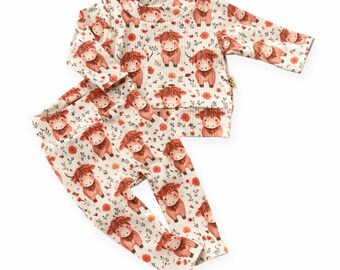 May include: A two-piece baby outfit featuring a cream-colored long-sleeved top and matching pants. The set is patterned with brown Highland cows and small orange flowers. The outfit is made of a soft, ribbed fabric.