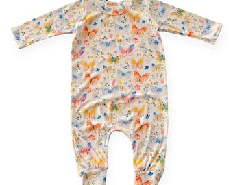 May include: A cream-colored baby romper with a ribbed texture and a colorful butterfly print. The long-sleeved one-piece features a round neckline and elastic cuffs. The butterflies are in shades of orange, yellow, blue, and pink.