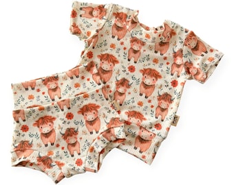 May include: A two-piece baby outfit featuring a cream-colored top and shorts. The set is patterned with cartoon Highland cows, red flowers, and green leaves. The top has short sleeves and the shorts have a high waist.