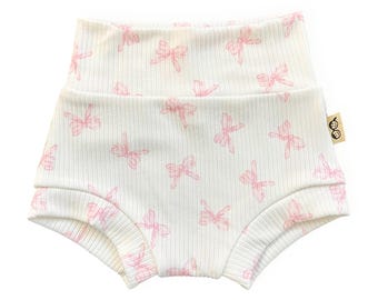 May include: White baby shorts with a pink bow pattern. The shorts have a high waist and are made of a ribbed fabric. The bows are evenly spaced across the shorts.