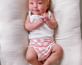 May include: A baby wearing a white tank top and pink and white checkered shorts. The baby is wearing a pink and white bow headband. The baby is lying on a white blanket.