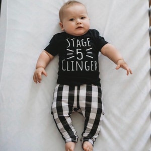 May include: A baby wearing a black t-shirt with the text "Stage 5 Clinger" in white. The baby is also wearing black and white checkered pants.