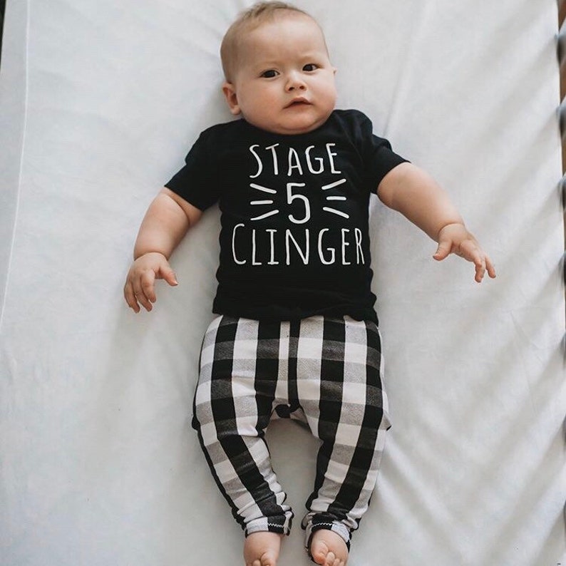 May include: A baby wearing a black t-shirt with the text "Stage 5 Clinger" in white. The baby is also wearing black and white checkered pants.