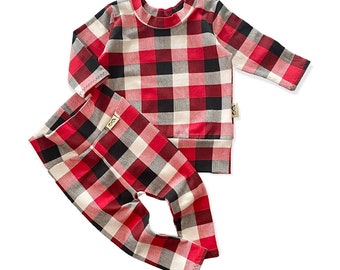 Op de afbeelding: Een tweedelige babyoutfit met een rood, zwart en wit ruitpatroon. De set bestaat uit een top met lange mouwen en een bijpassende broek. De top heeft een ronde hals en de broek is nauwsluitend.