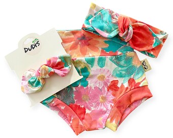 May include: A floral print baby set featuring a pair of bloomers, a headband, and a matching bow. The set has a white background with pink, orange, and teal flowers. The brand name "DUDIS" is visible on the tag.