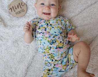 May include: A baby wearing a floral print outfit and matching headband lies on a fluffy white blanket. A wooden disc with the words "nine months" is next to the baby. The outfit features blue, pink, and yellow flowers.
