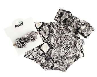 May include: A black and white snake print baby headband and matching bib. The headband is tied in a bow and the bib has a small tag with the word "DUDIS" on it.