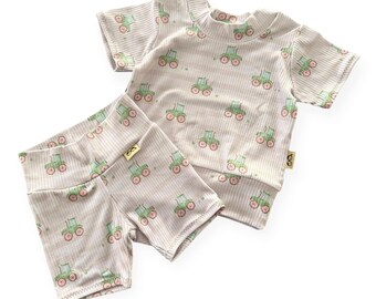 May include: A two-piece baby outfit featuring a short-sleeved shirt and shorts. The set is light beige with a ribbed texture and a repeating pattern of green tractors with pink wheels. The outfit is designed for comfort and style.