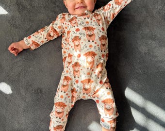 May include: A baby wearing a cream-colored, long-sleeved romper with a repeating pattern of brown Highland cows and orange floral accents. The romper has a ribbed texture and is laying on a gray surface.