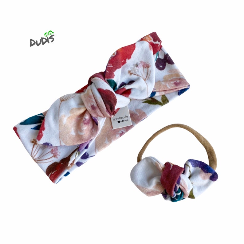 May include: A set of two floral print headbands. The larger headband is tied in a bow and has a white background with red, pink, and purple flowers. The smaller headband is a nude elastic with a matching floral print bow. The tag on the larger headband reads "Handmade with Love".