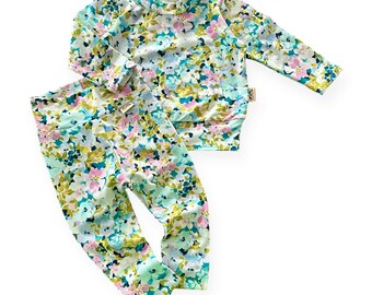 May include: A two-piece floral set featuring a long-sleeved top and leggings. The set is adorned with a delicate floral pattern in shades of blue, pink, yellow, and green on a light background. The top has a mock neck, and the leggings have a high waist.