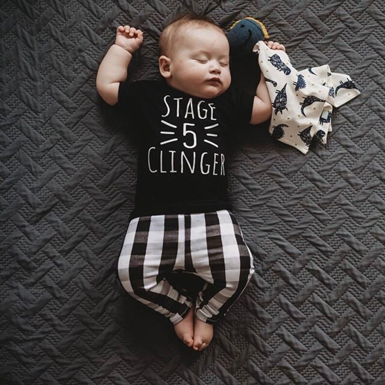 May include: A baby wearing a black t-shirt with the text "Stage 5 Clinger" in white. The baby is wearing black and white checkered pants and is lying on a gray quilted blanket.