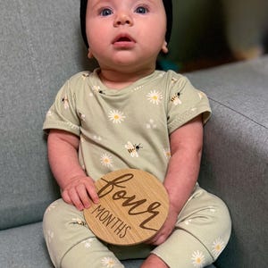 Pode incluir: Um bebé com um conjunto verde claro com um padrão de margaridas e abelhas, e uma faixa preta com laço. O bebé segura um disco de madeira com a inscrição "four MONTHS". O bebé está sentado num sofá cinzento.