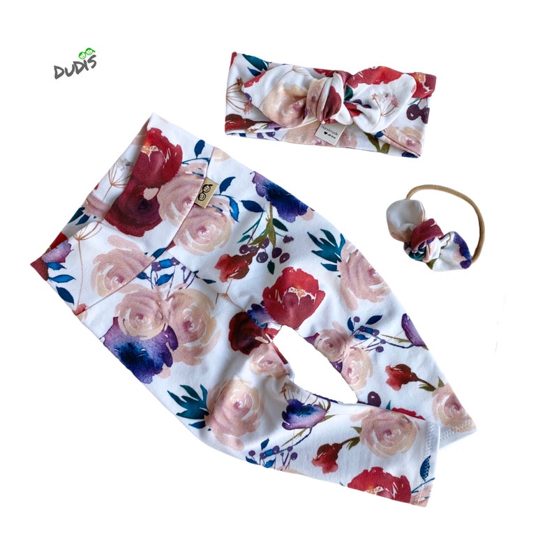 May include: A set of baby leggings and two headbands with a floral print. The leggings are white with a watercolor floral pattern in shades of pink, red, purple, and green. The headbands are a light brown color with a floral print.