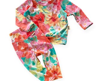 May include: A two-piece floral outfit featuring a long-sleeved top and leggings. The set showcases a vibrant pattern of red, pink, yellow, and teal flowers. The fabric appears soft and comfortable, suitable for casual wear.