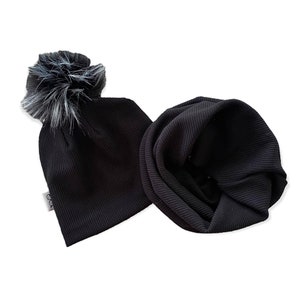 May include: A black ribbed knit beanie with a faux fur pom-pom and a matching black ribbed knit infinity scarf. The beanie has a small tag on the side. The set is displayed on a white background.