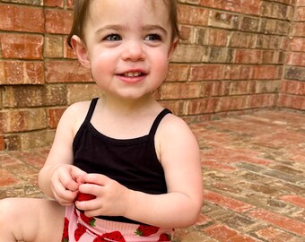 May include: A black spaghetti strap tank top paired with pink shorts featuring a red strawberry print. A matching pink strawberry print scrunchie is worn in the hair. The outfit is made of a ribbed fabric and includes a small white tag on the shorts.