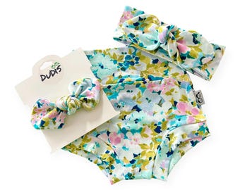 May include: A baby clothing set featuring a floral print in shades of blue, pink, and green. The set includes a pair of bloomers, a headband, and a bow. The brand name "DUDIS" is printed on the tag.