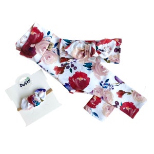 May include: A set of three floral patterned baby accessories: a headband, leggings, and a hair bow. The headband and leggings feature a white background with red, pink, and purple watercolor flowers. The hair bow is a similar floral pattern with a brown elastic band.
