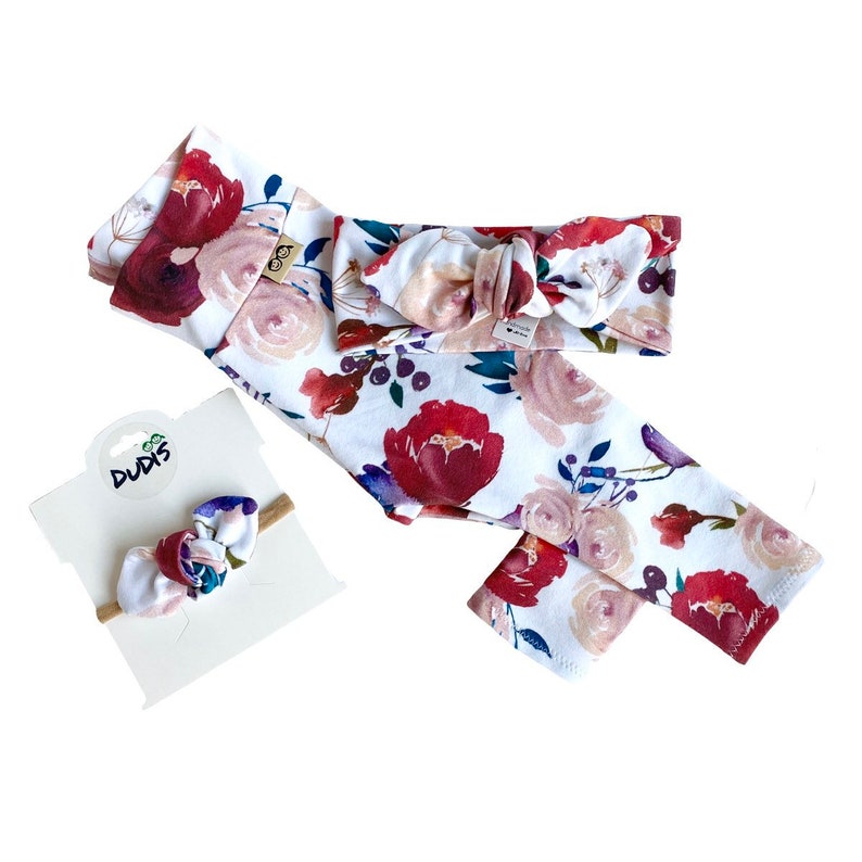 May include: A set of three floral patterned baby accessories: a headband, leggings, and a hair bow. The headband and leggings feature a white background with red, pink, and purple watercolor flowers. The hair bow is a similar floral pattern with a brown elastic band.