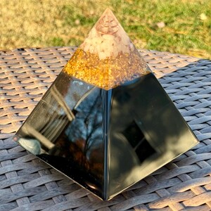 May include: A decorative pyramid with a black base, a gold-colored middle section, and a clear top with a copper coil. The pyramid is sitting on a brown woven surface, with a blurred green background.