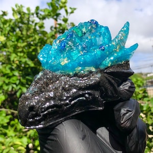 May include: A black and blue resin dragon head sculpture with glitter embedded in the resin. The dragon's head is facing the left side of the image. The dragon's head is covered in a blue resin with gold glitter embedded in the resin. The dragon's head is sitting on a black surface.