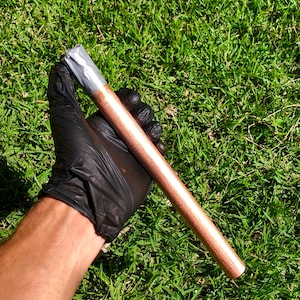 Puede incluir: Un tubo de color cobre con cinta plateada envuelta alrededor de un extremo. El tubo está siendo sostenido por una mano enguantada.