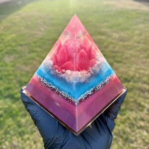 May include: A decorative pyramid with a layered design. The base is a deep purple, transitioning to blue, and topped with a pink floral arrangement. Silver glitter accents the layers. The pyramid is held in a black gloved hand.
