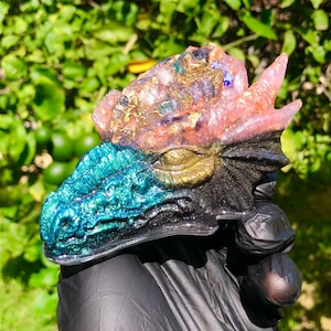 May include: A resin dragon head sculpture with a pink, gold, and blue glitter finish. The dragon's eye is gold and the scales are a teal and black glitter.