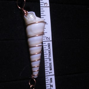Puede incluir: Un colgante de concha blanca con alambre de cobre envuelto alrededor. El colgante mide aproximadamente 5 cm de largo y tiene una perla en la parte inferior. El colgante cuelga de un cordón negro y está al lado de una regla.